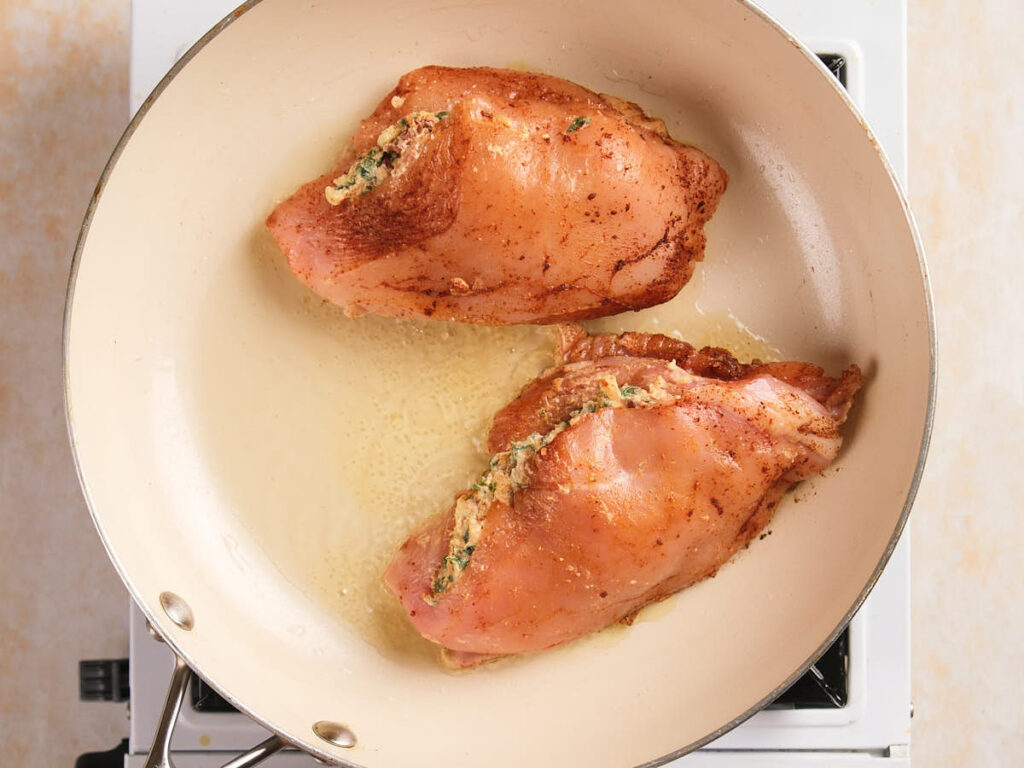 Two stuffed chicken breasts cooking in a cream-colored skillet on a stovetop.