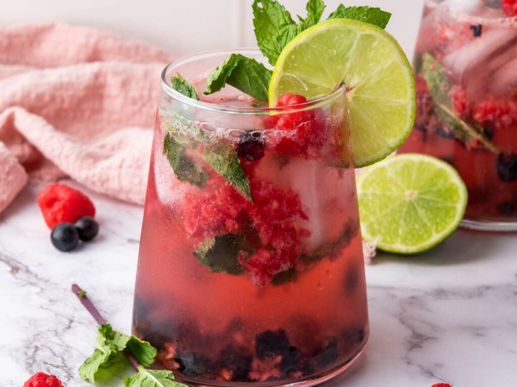 A glass of blueberry mocktail with fresh mint and lime, a pink hand towel visible in the background.