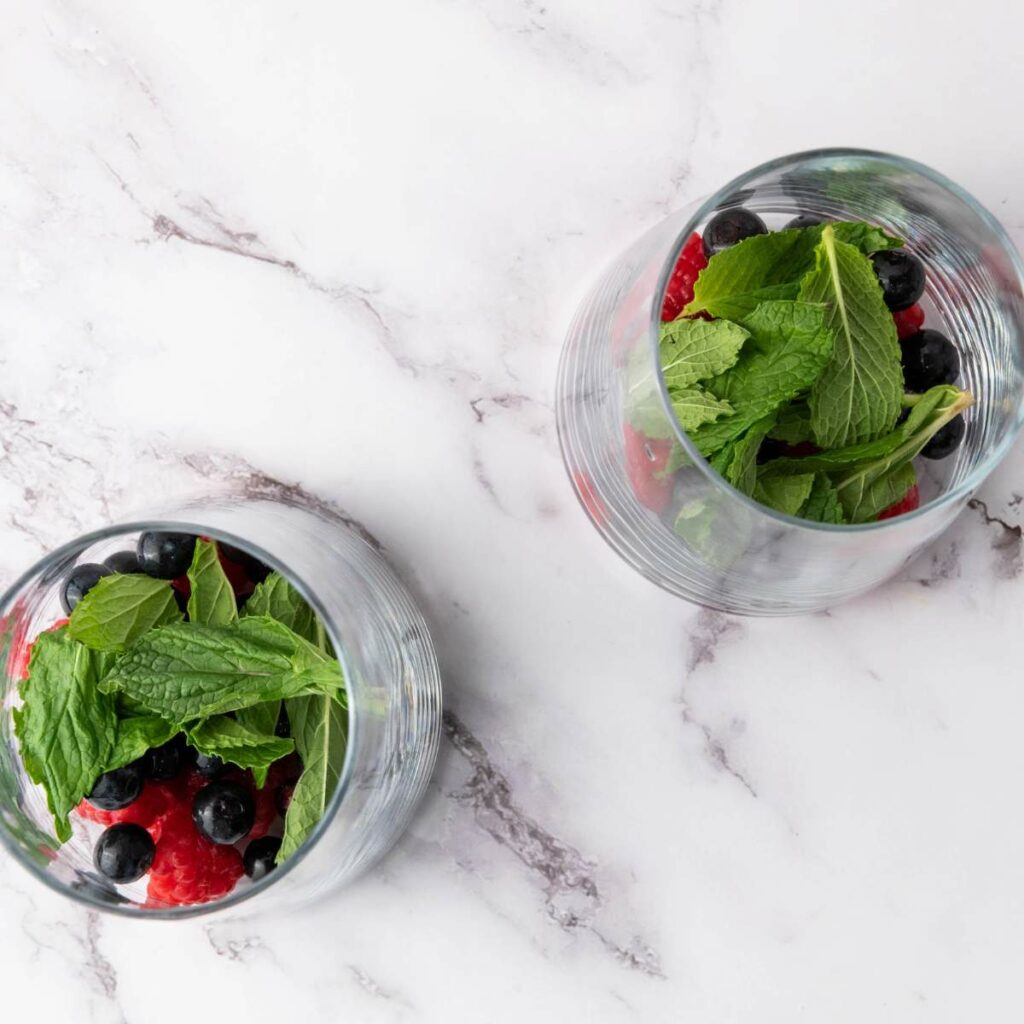 Two glasses with blueberries, raspberries and mint leaves on a white marble surface.