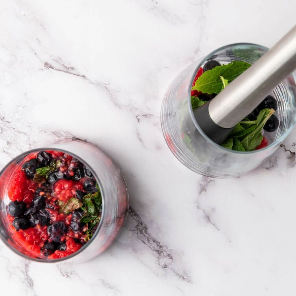 Two glasses on a marble surface, one filled with crushed berries and herbs, the other with fresh mint and a muddler.