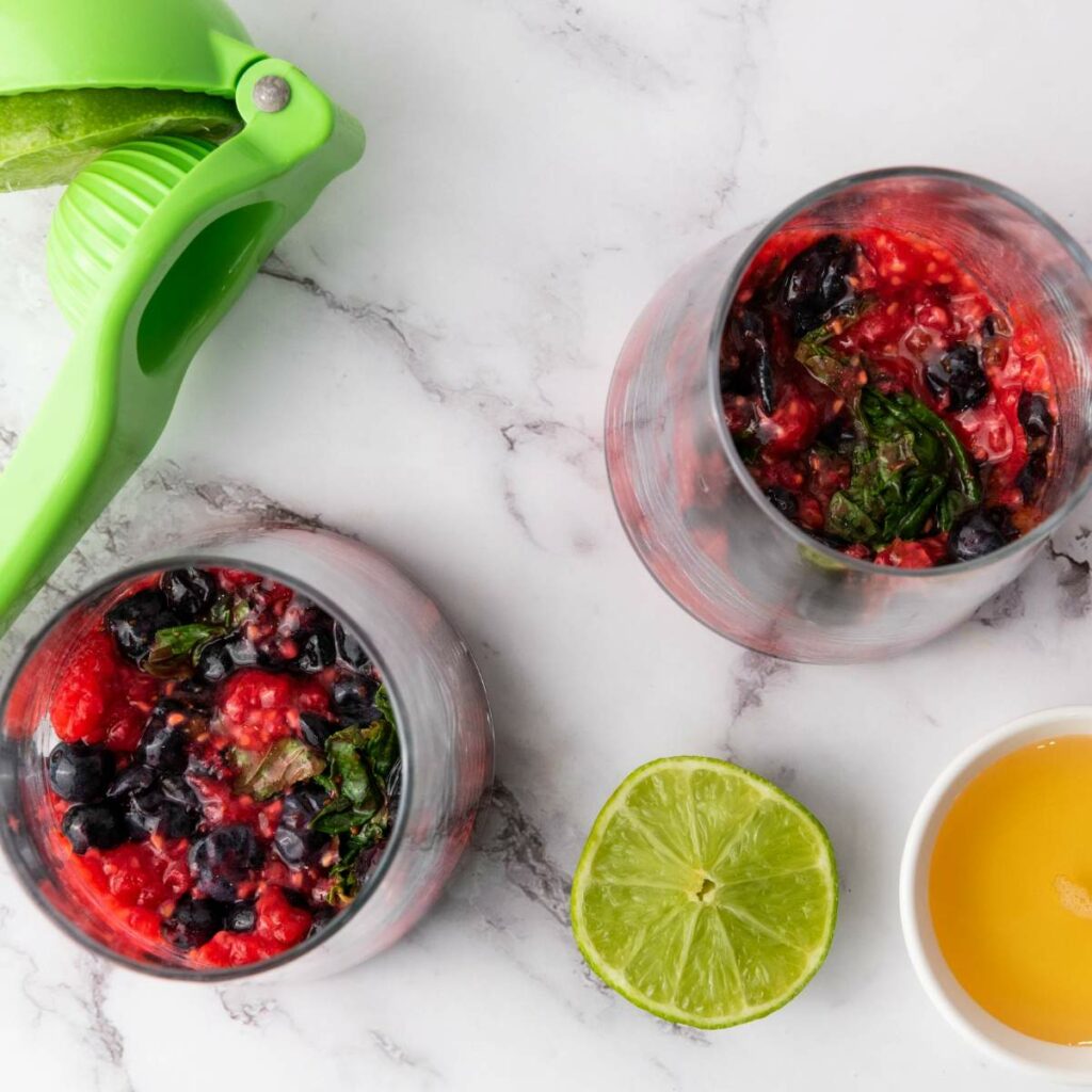 Two glasses filled with a berry and mint mixture alongside a green citrus squeezer, half a lime, and a small bowl of liquid sweeter.