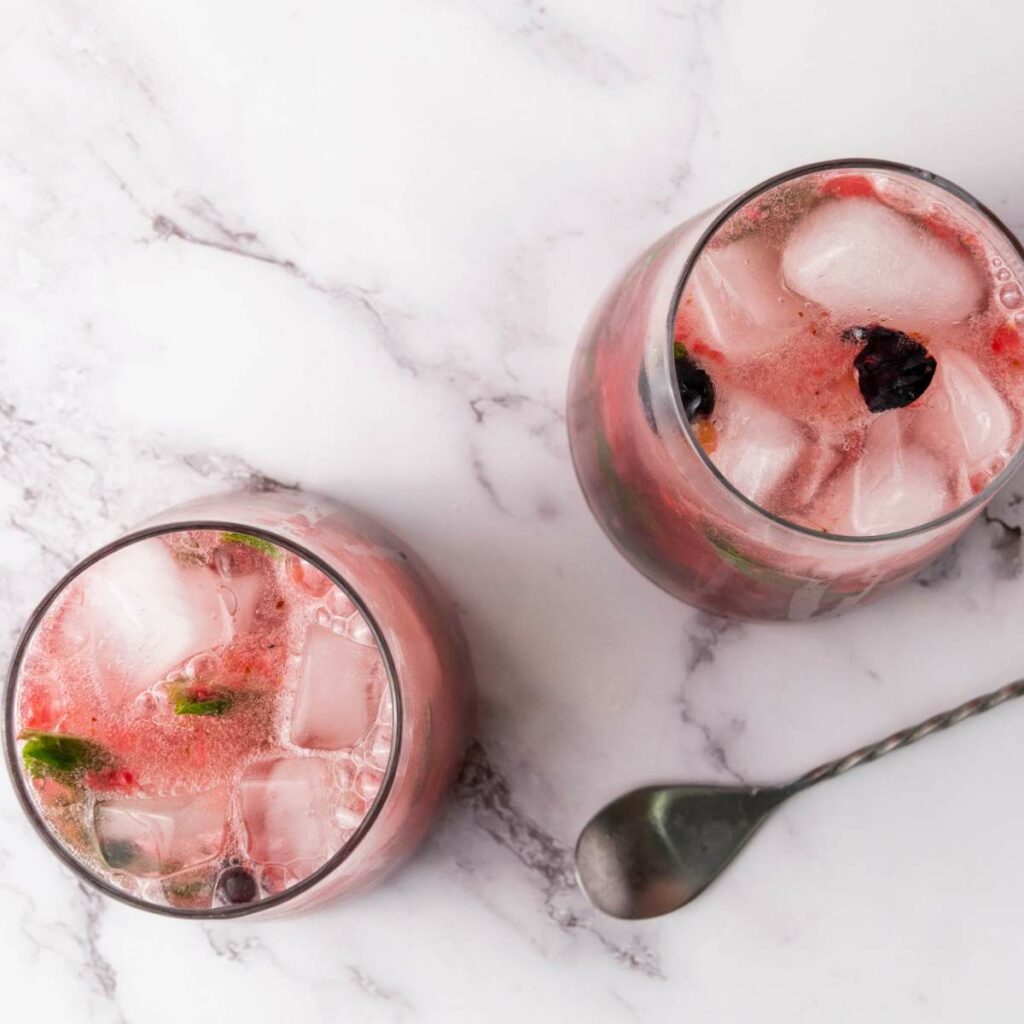Two glasses filled with a pink berry mocktail and ice sit on a marble surface beside a metal spoon.