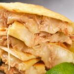 Close-up of stacked chicken quesadillas filled with melted cheese and shredded chicken, with a lime wedge in the foreground.