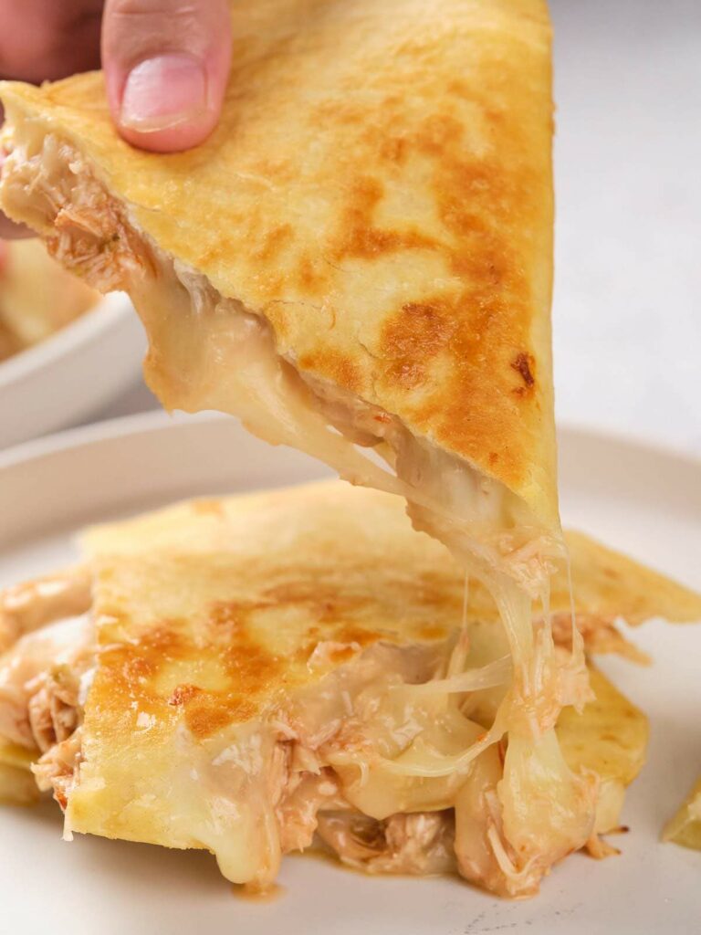 A hand lifts a cheesy chicken quesadilla slice from a plate, showing melted cheese stretching on a white plate.