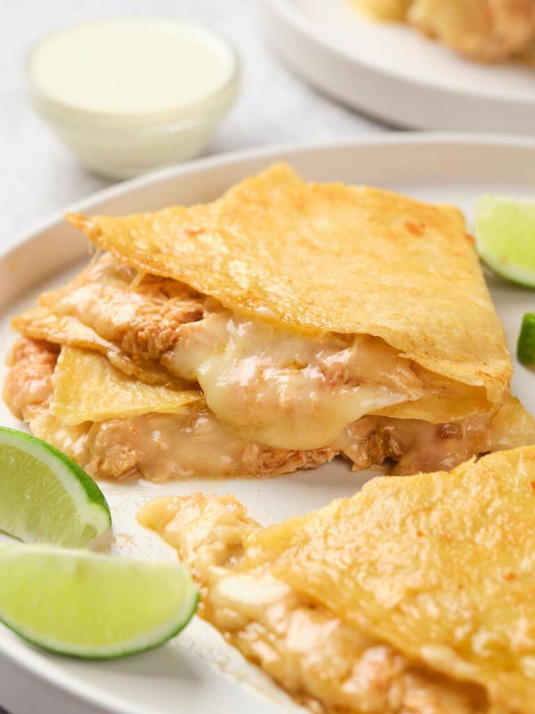 A white plate of chicken quesadillas garnished with lime wedges, with a small bowl of sauce in the background.