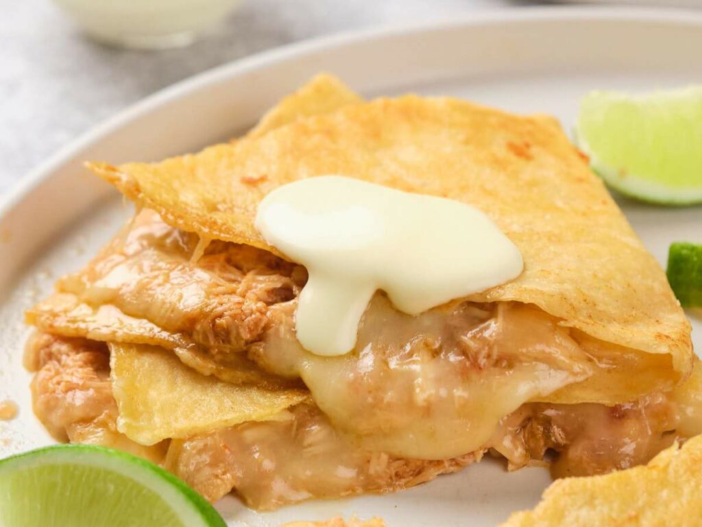 A close-up of a chicken quesadillas with white cream on top, served on a white plate with lime wedges.
