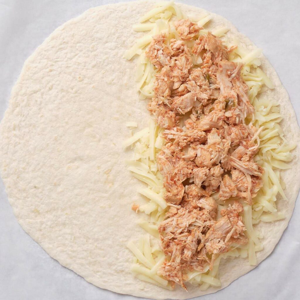 A tortilla with shredded cheese and shredded chicken arranged on one half, ready to be folded.