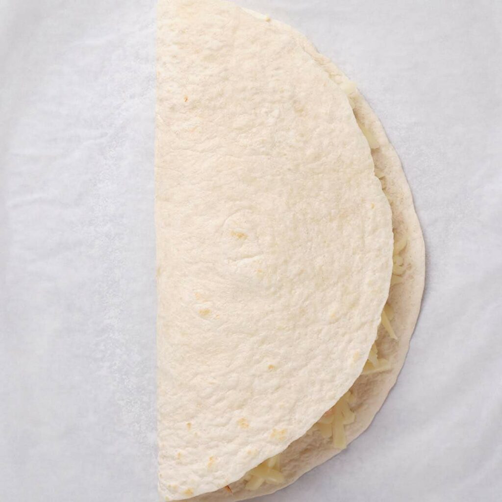 A flour tortilla folded in half with shredded cheese and shredded chicken inside, placed on a white surface.