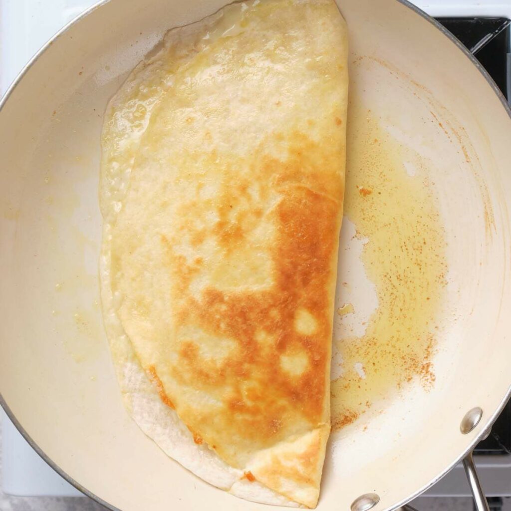 A folded quesadilla in half is being cooked in a lightly oiled white pan on a stovetop.