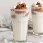 A glasses of milk shake with mini marshmallow on a marble surface next to a striped table cloth on the background.