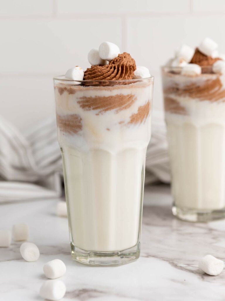 A glasses of chocolate milk with mini marshmallow on a marble surface next to a striped table cloth on the background.