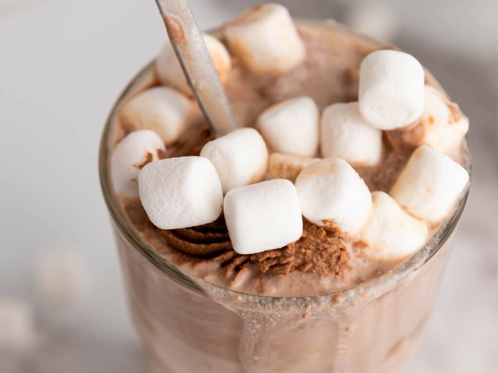 A chocolate milk filled with chocolate whipped cream and mini marshmallows with a spoon inside.