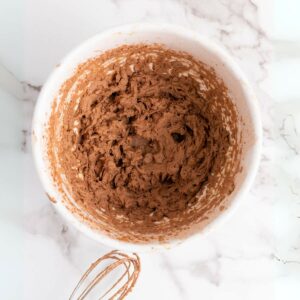 A white bowl of a mixture of chocolate cream next to a hand mixer on a white marble surface.