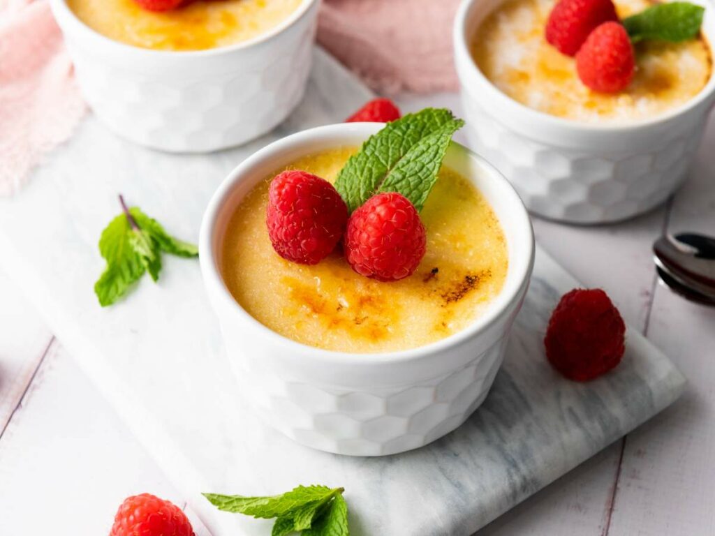A white ramekin of creme brulee topped with two raspberries and a sprig of mint.
