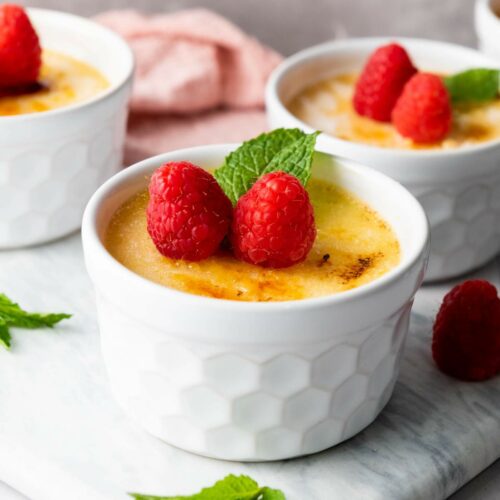 A white ramekin with crème brûlée topped with two raspberries and a mint leaf, placed on a marble surface, with more ramekins and raspberries in the background.