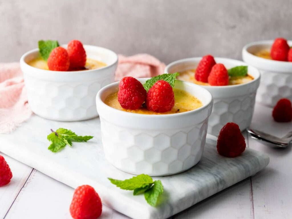 Four white ramekins filled with creme brulee, each topped with fresh raspberries and mint leaves.