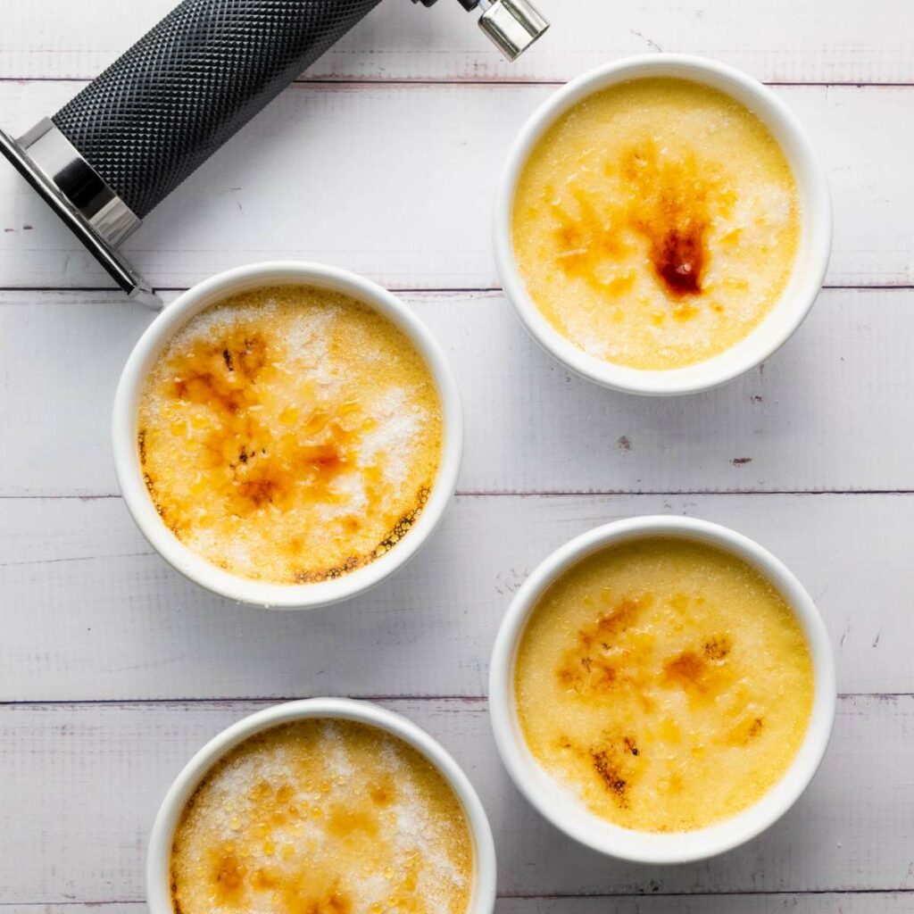 Four ramekins of crème brûlée with caramelized tops on a white wooden surface.