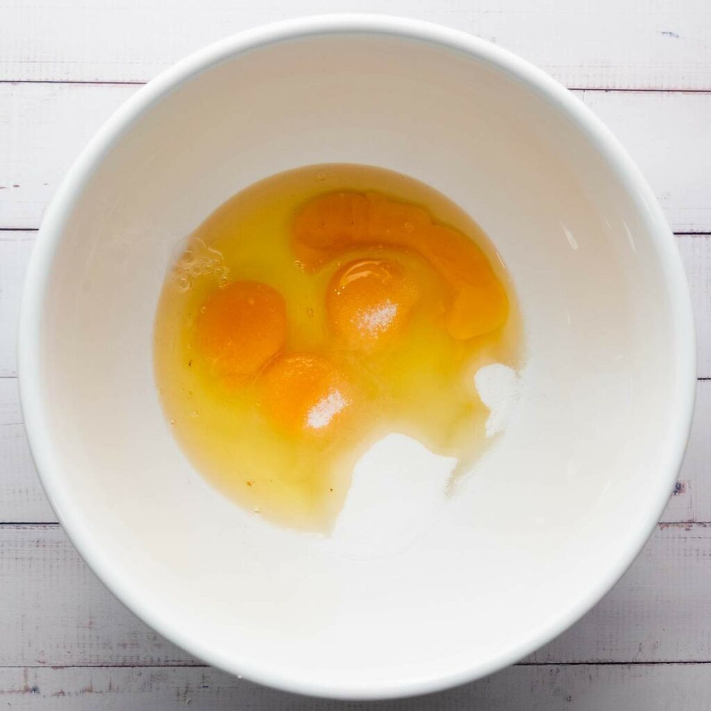Three raw eggs cracked into a large white bowl on a white wooden surface.