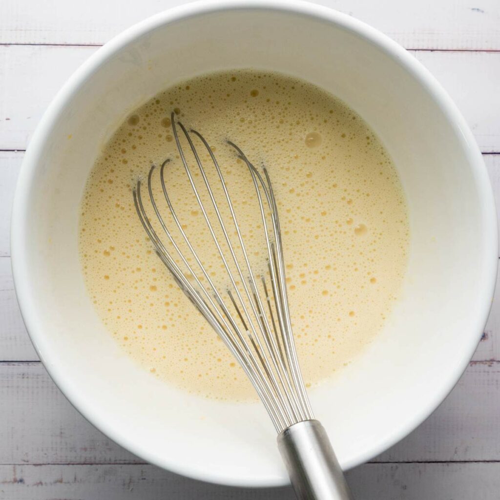 A metal whisk rests in a white bowl filled with a light yellow, frothy batter.