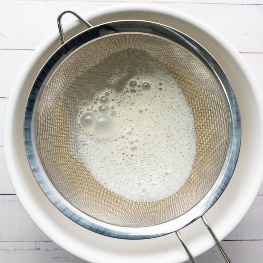 A metal strainer with frothy liquid rests over a white bowl.