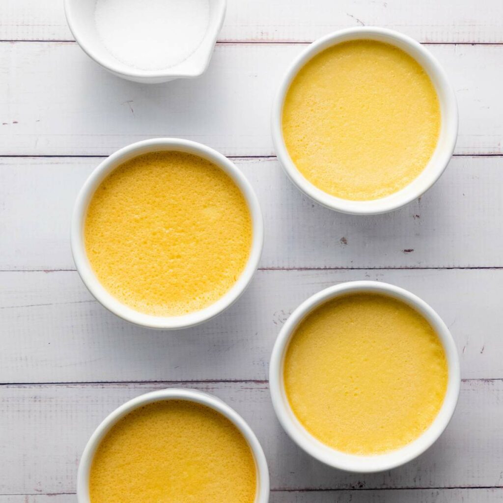 Four white ramekins filled with yellow custard sit on a white wooden surface.