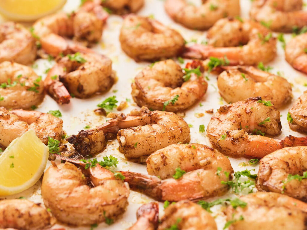 Garlic butter shrimp seasoned with spices and garnished with chopped parsley, arranged on a tray with lemon wedges.