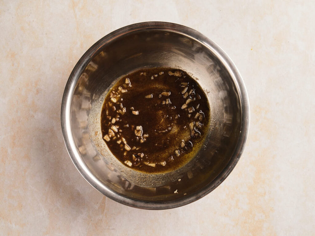 A stainless steel bowl containing a mixture of chopped garlic and a brown liquid marinade.