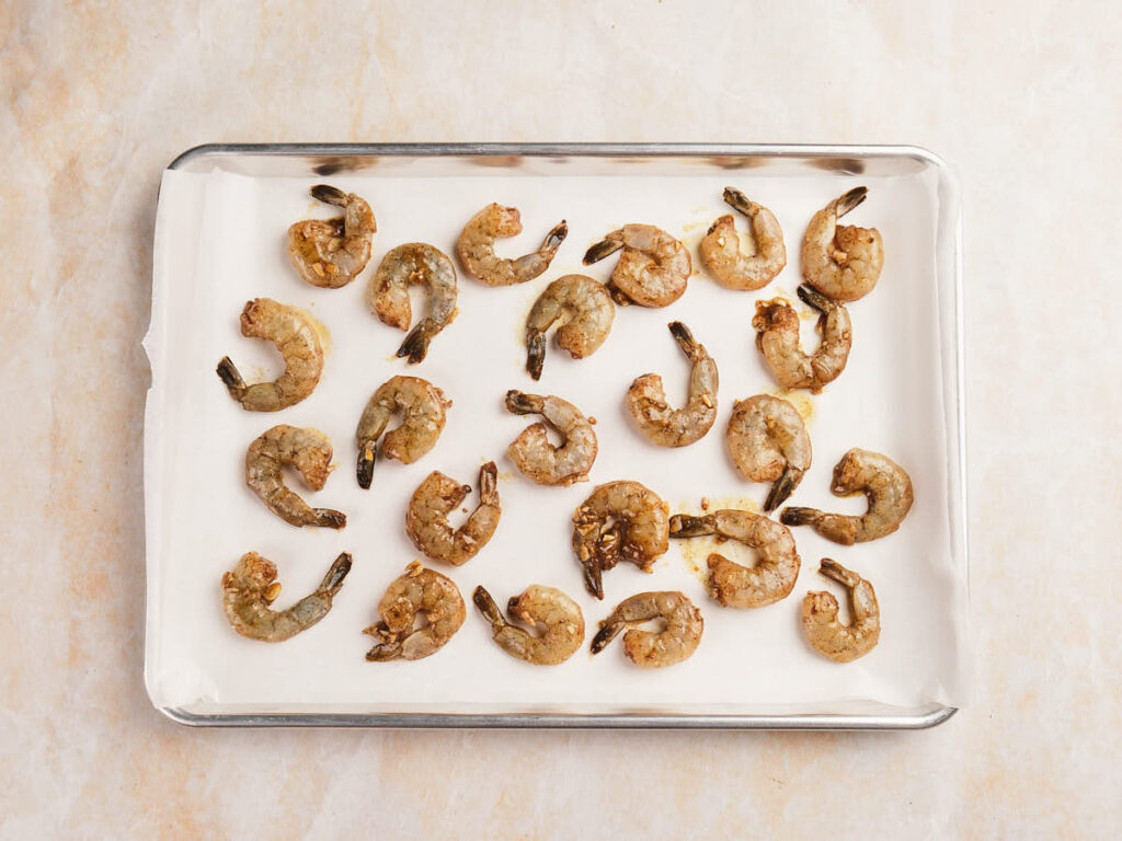 Raw marinated shrimp arranged in rows on a parchment-lined baking sheet.