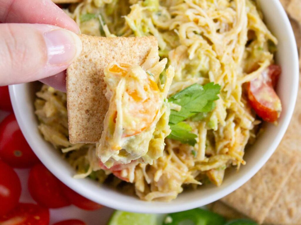 A hand holds a cracker topped with a healthy chicken salad and vegetable dip.