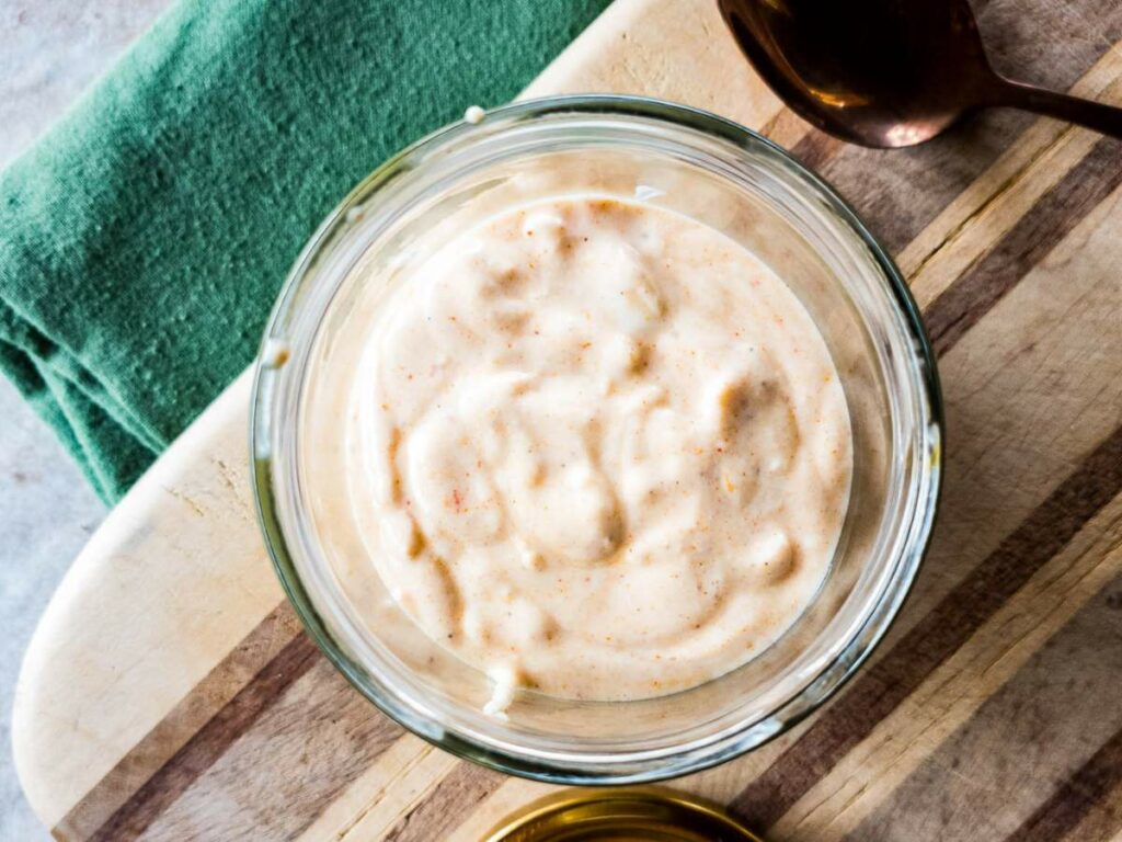 A glass bowl of creamy horseradish sauce sits on a wooden surface next to a dark green towel.