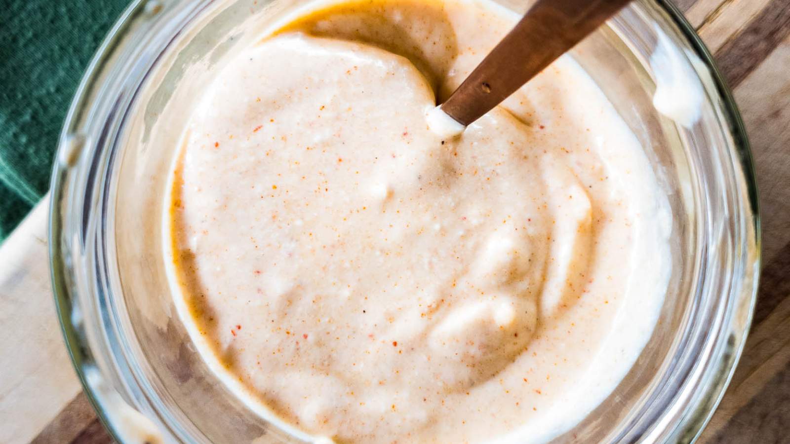 A glass bowl filled with creamy horseradish sauce is shown with a spoon on a wooden board.