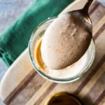 A spoonful of horseradish sauce is held above a glass bowl on a wooden board and a dark green towel next to it.