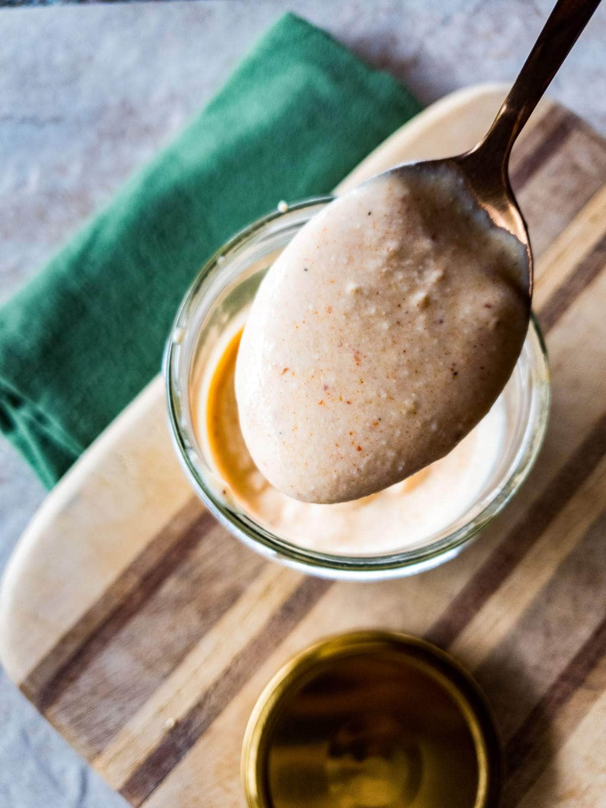 A spoonful of horseradish sauce is held above a glass bowl on a wooden board and a dark green towel next to it.