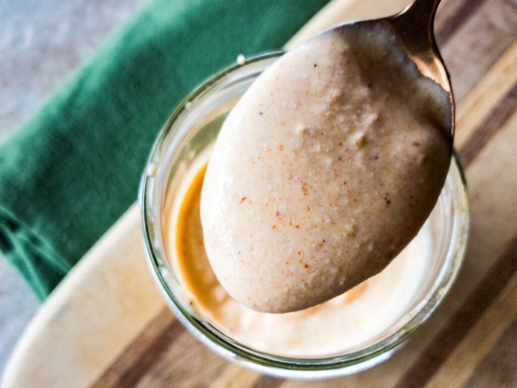 A spoonful of creamy horseradish sauce is held above a glass bowl on a wooden surface.