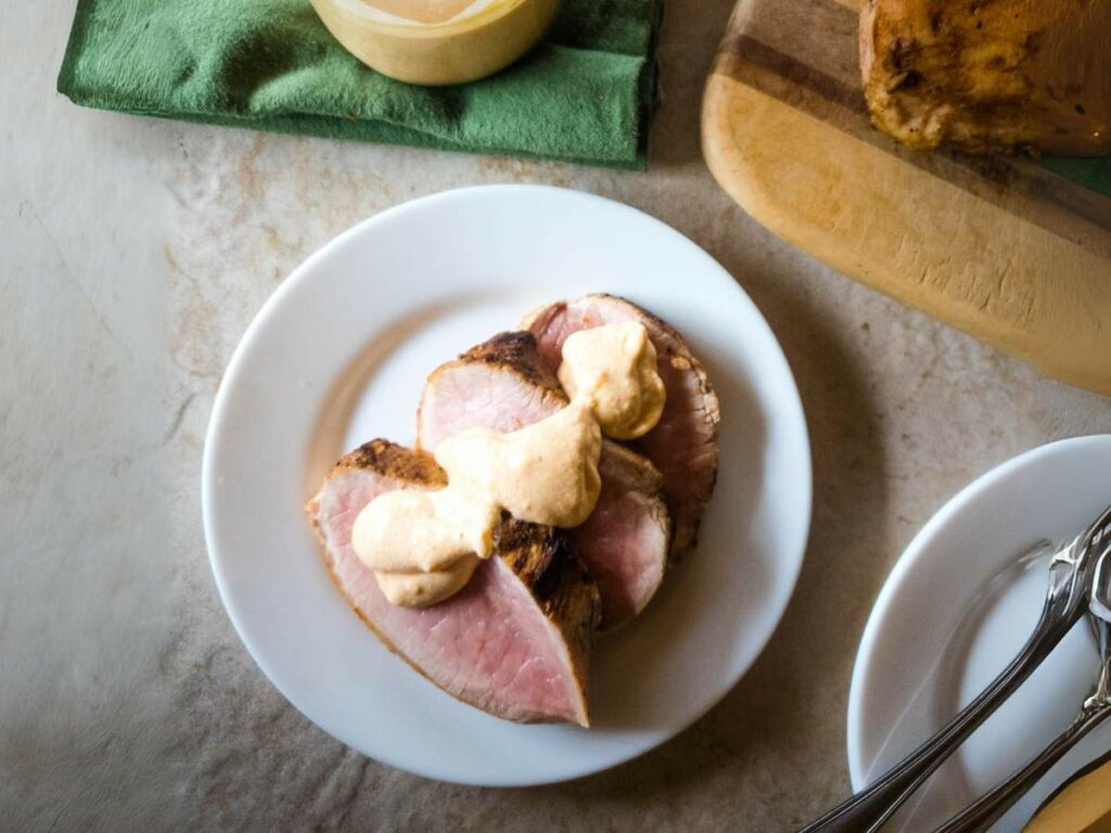 A white plate of three sliced meat topped with horseradish sauce on a white surface.