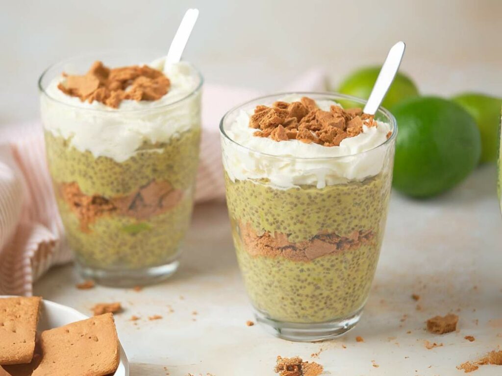 Two glasses of chia pudding topped with cream and crackers sitting on a white surface.