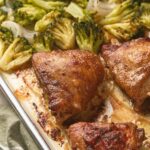 Baked chicken thighs with broccoli florets and onions on a single baking sheet.