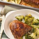 A plate with a piece of sheet pan chicken thighs and a side of cooked broccoli and onions.