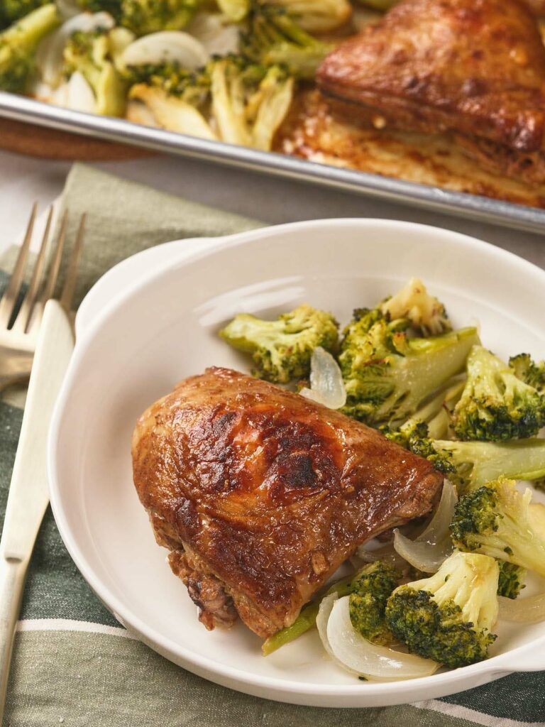 A plate with a piece of sheet pan chicken thighs and a side of cooked broccoli and onions.