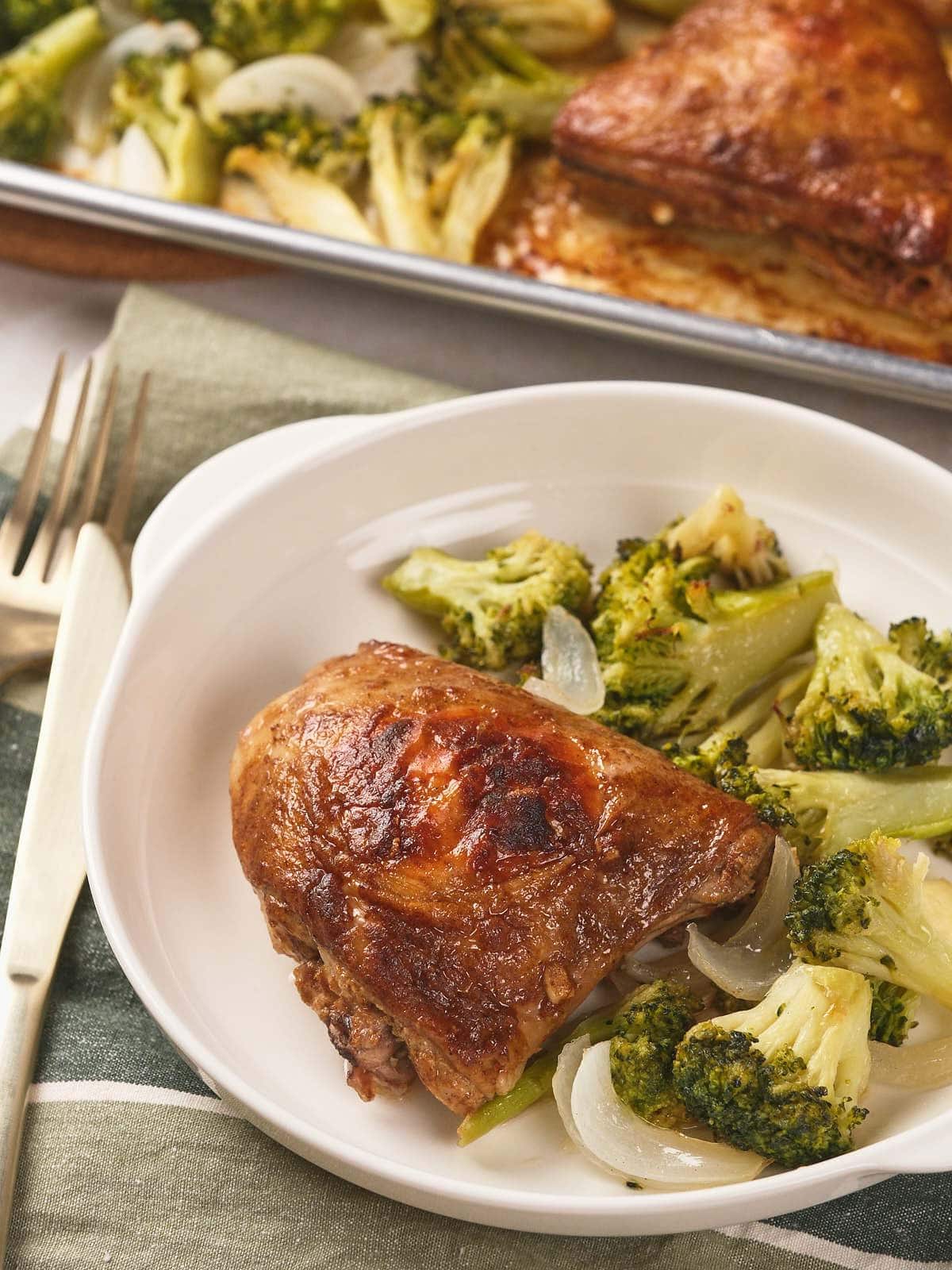 A plate with a piece of sheet pan chicken thighs and a side of cooked broccoli and onions.