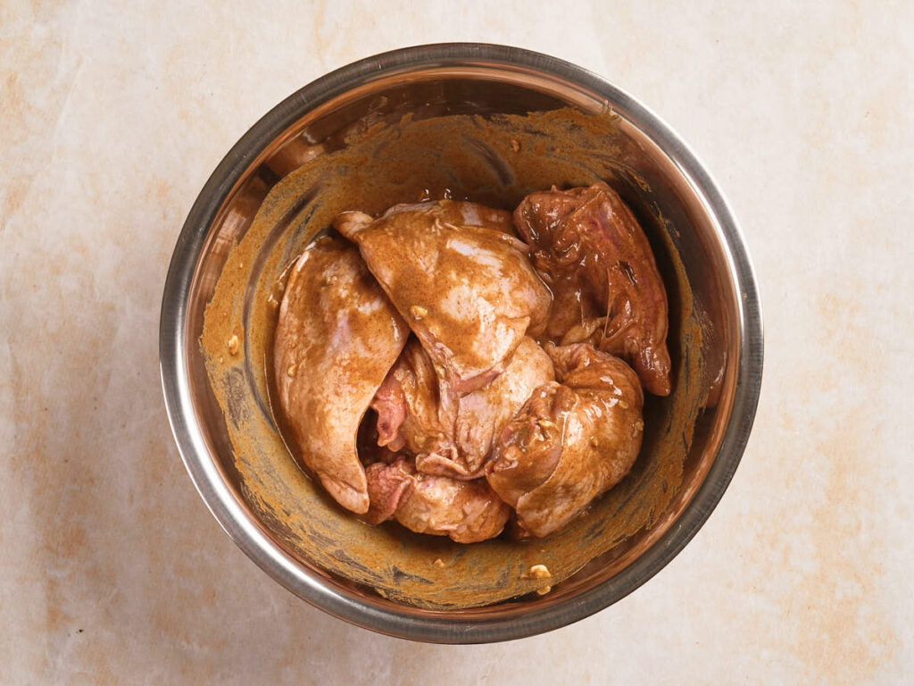 Raw chicken pieces coated in a brown marinade are placed in a stainless steel mixing bowl.