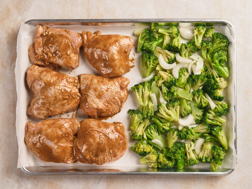 Sheet pan with four marinated chicken thighs on one side and a mixture of chopped broccoli.
