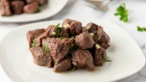 A white plate with cooked steak garnished with chopped herbs, with another plate and some parsley in the background.