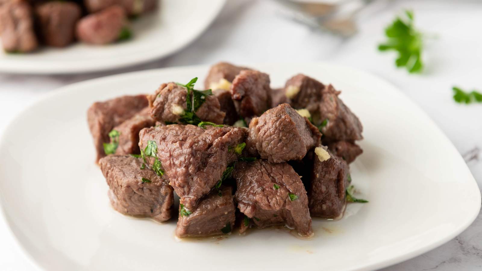 A white plate with cooked steak bites garnished with chopped herbs, with another plate and some parsley in the background.