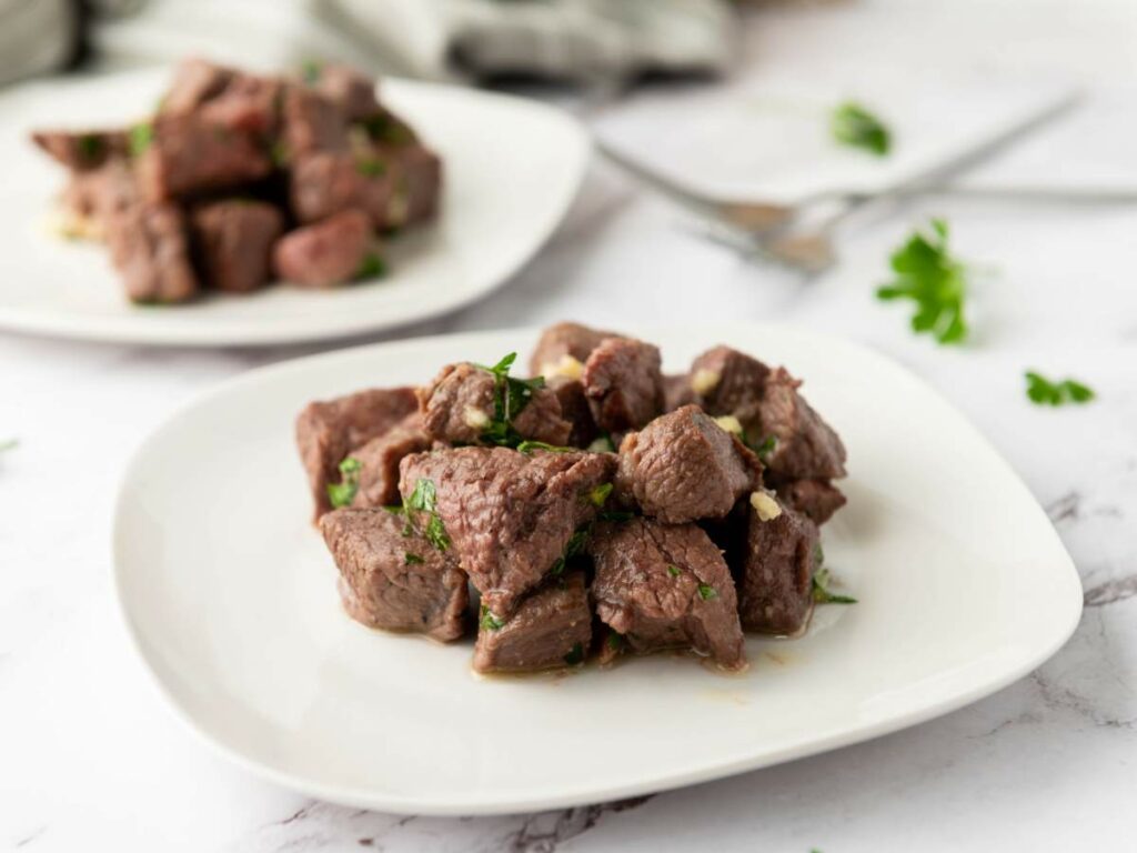 Cubes of cooked steak bites garnished with herbs are served on a white plate.