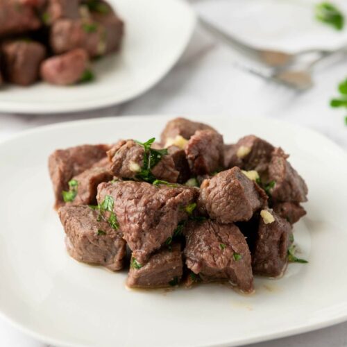 Cubed steak bites garnished with chopped herbs are arranged on a white plate, with another plate of beef cubes visible in the background.