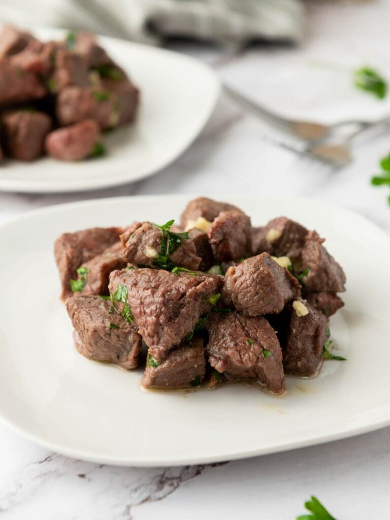 Cubed steak bites garnished with chopped herbs are arranged on a white plate, with another plate of beef cubes visible in the background.