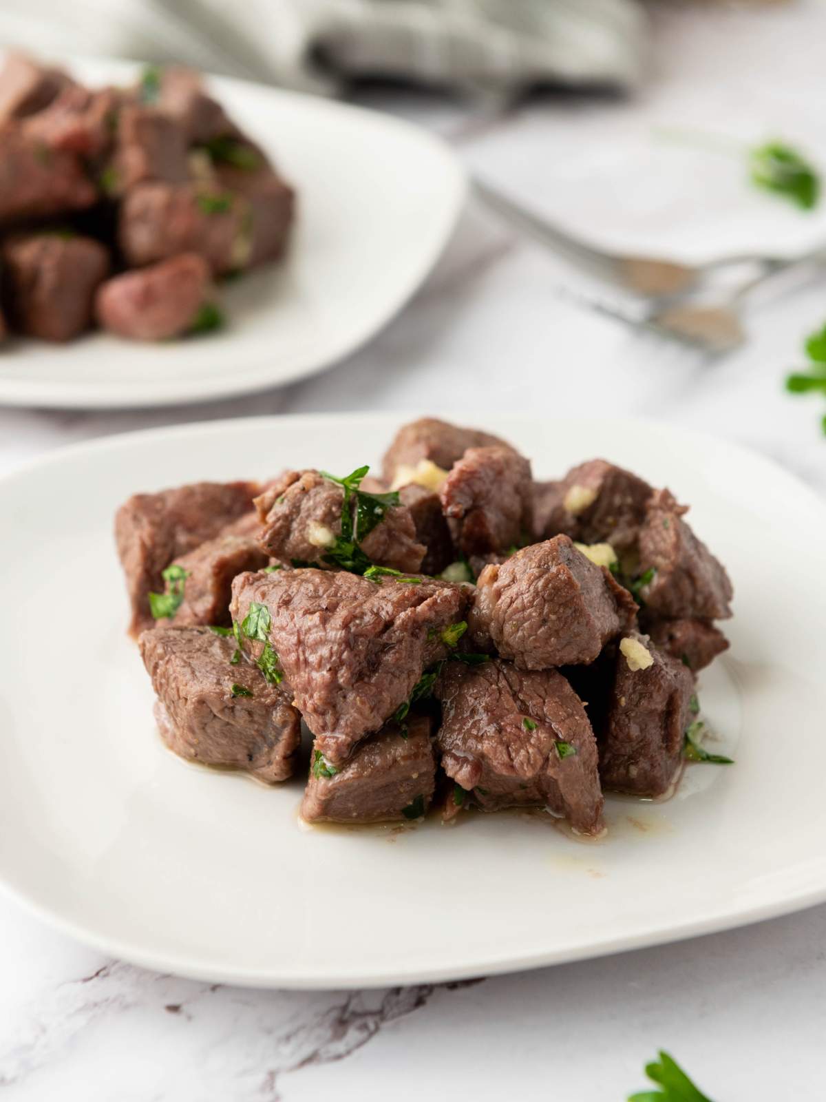 Cubed steak bites garnished with chopped herbs are arranged on a white plate, with another plate of beef cubes visible in the background.