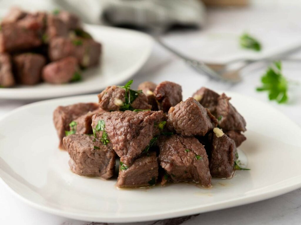 A plate of cooked steak bites cubes garnished with chopped herbs.
