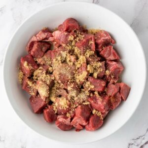 Cubed raw steak bites in a white bowl, topped with a layer of brown sugar and spices, on a white marble surface.